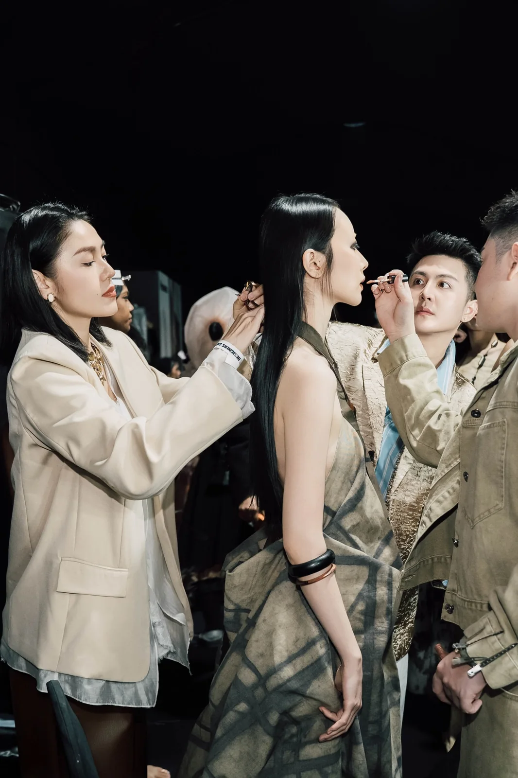 Backstage before a show: a make-up artist in a white blazer applies lip colour to a model while another artist works beside them