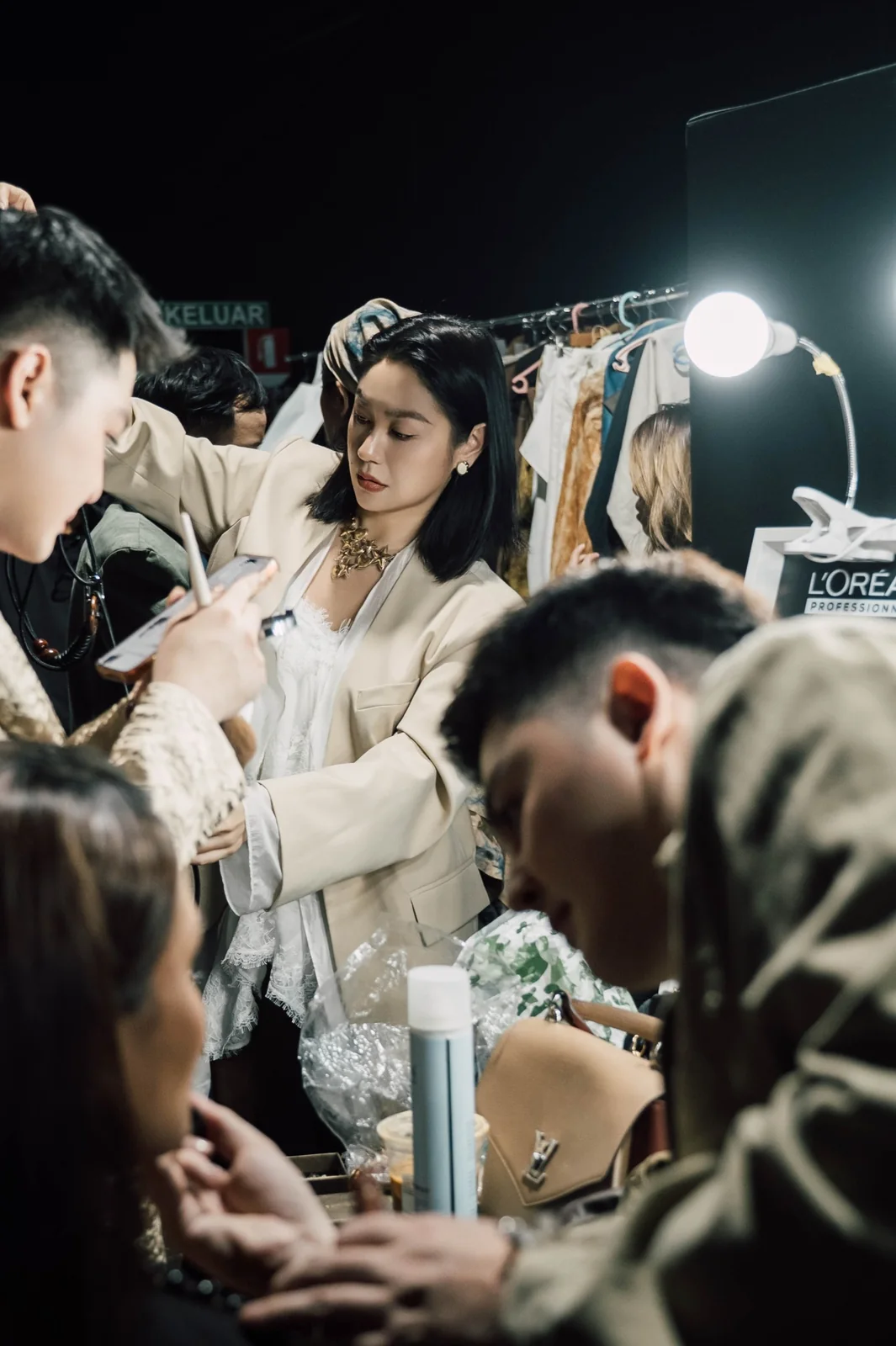 Backstage at a show: a stylist in a white blazer works beside a clothing rail while a make-up artist works in the foreground
