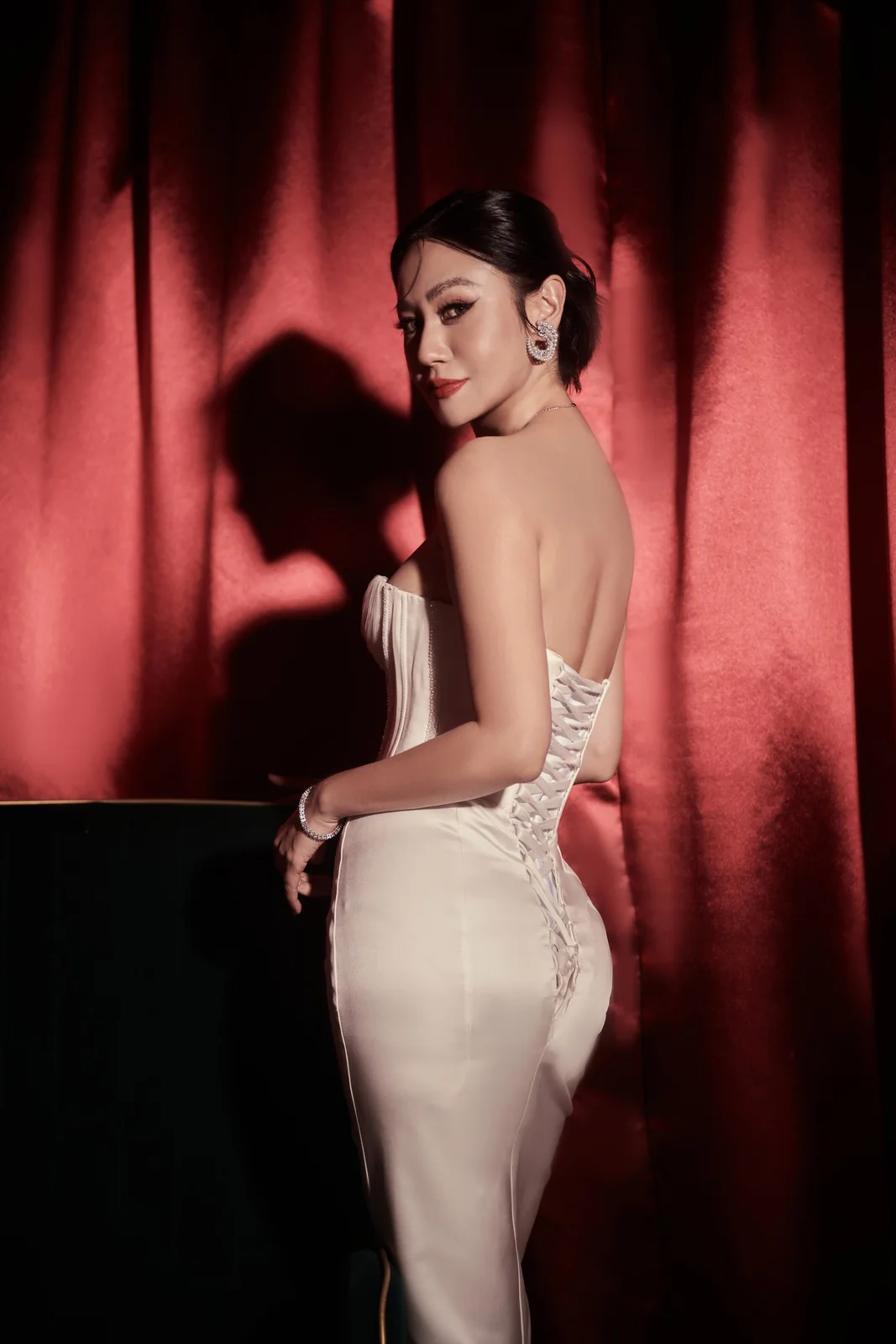 Goey in an ivory corseted gown against a dark curtain, looking back over her shoulder
