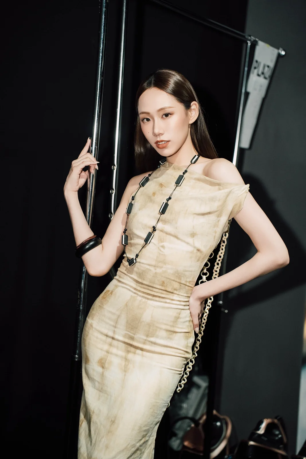 Nico backstage in a draped ivory dress and a long bead necklace, one hand on a clothing rail