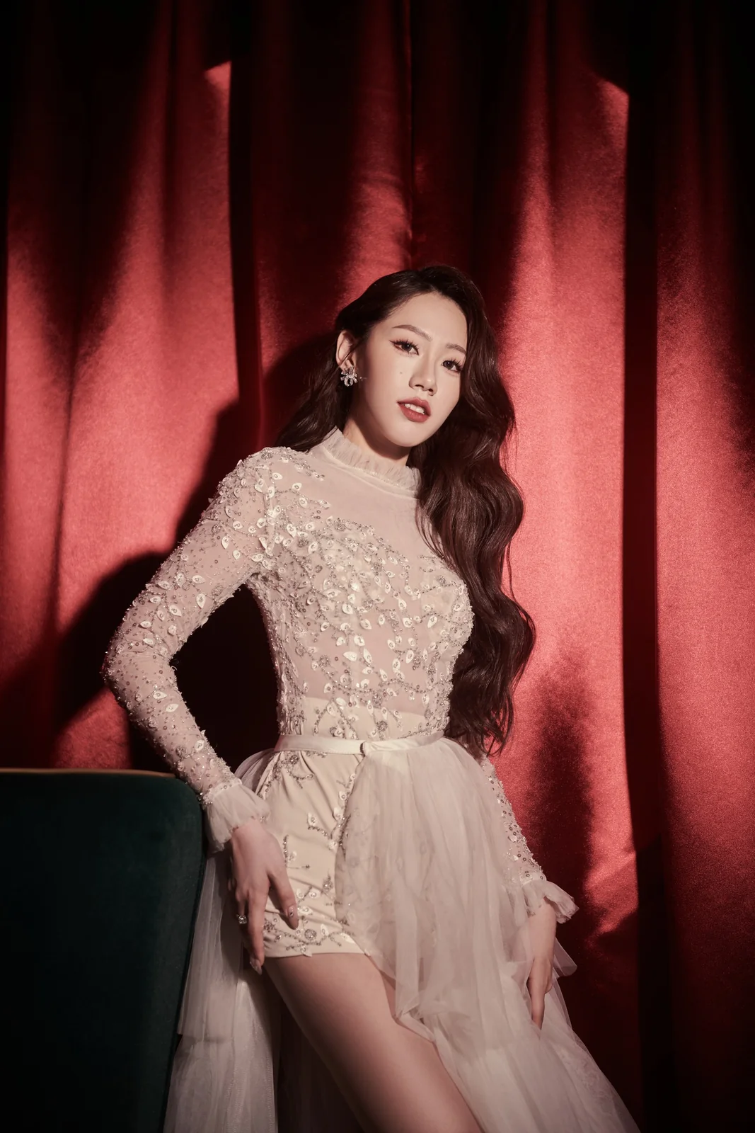 Editorial portrait: Nico in a long-sleeved lace gown with a sheer skirt, seated before a dark curtain