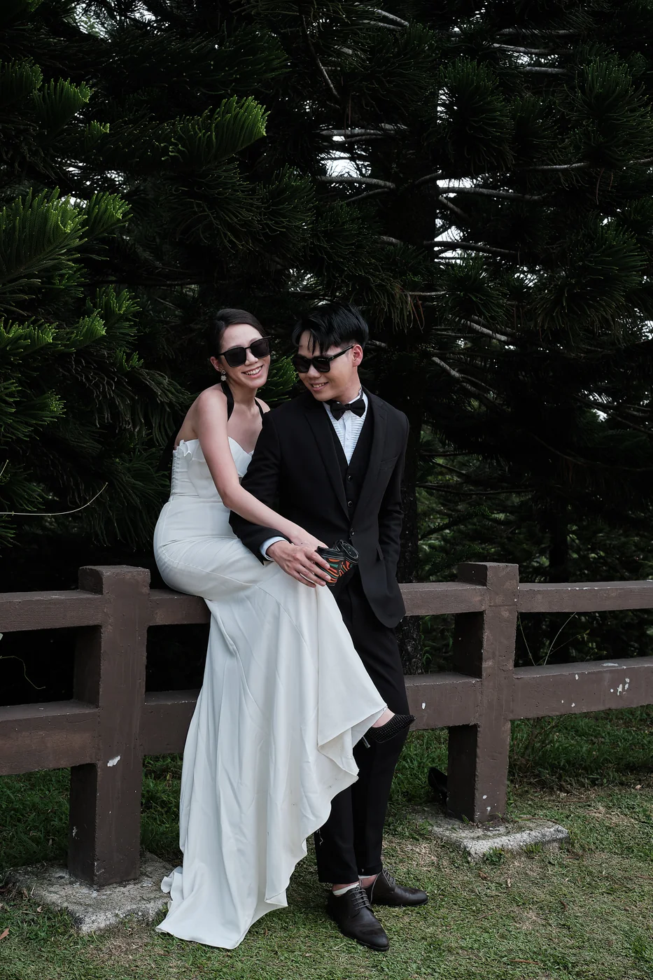 Pre-wedding shoot: the couple in sunglasses pose against a wooden fence, the bride in a white gown