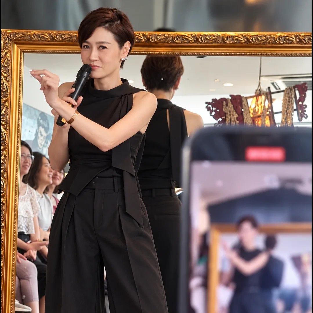 A teacher with a microphone demonstrates a gesture in front of a large gilded mirror, a class watching