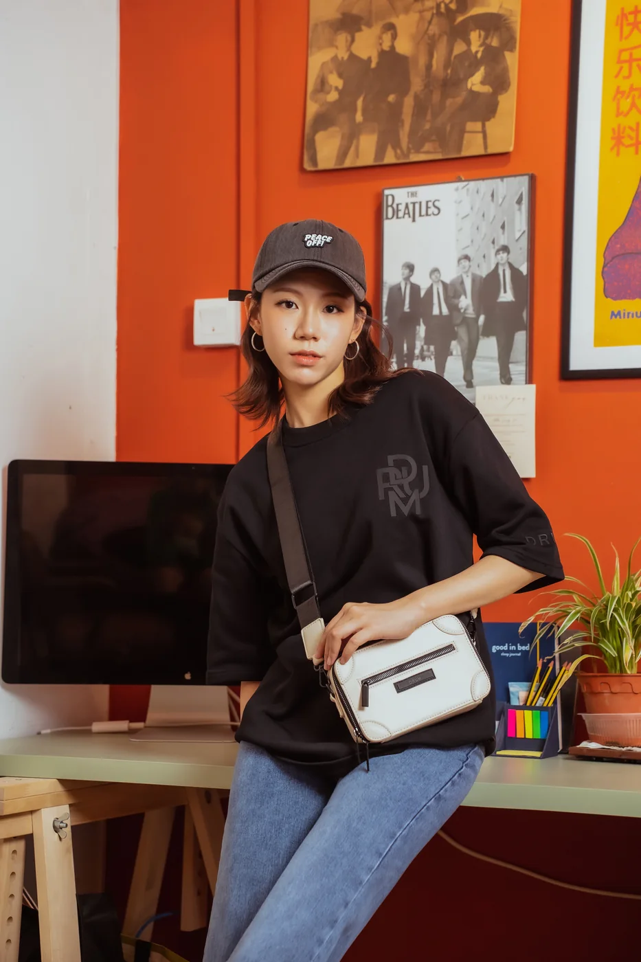 Lifestyle shoot: a model in a cap and an oversized T-shirt with a crossbody bag, leaning on a desk