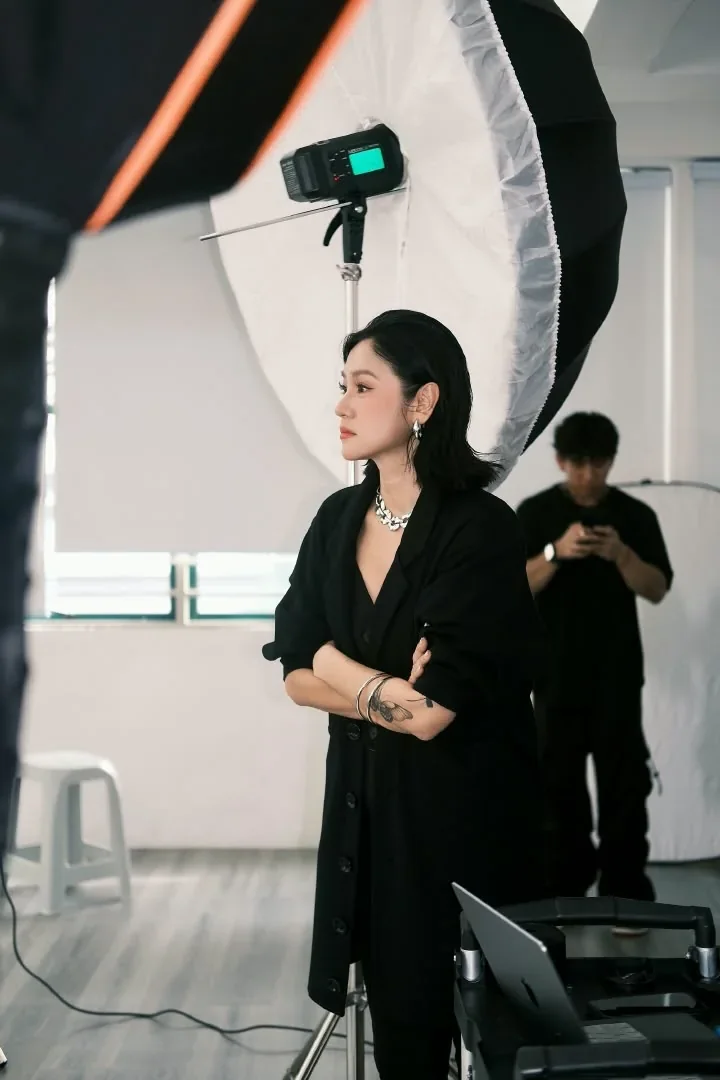 Directing a studio shoot beside a large softbox, arms folded, a photographer behind