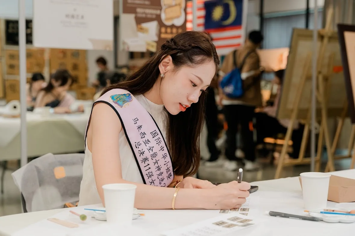 A contestant wearing a pageant sash writes at a table, easels and other contestants behind her
