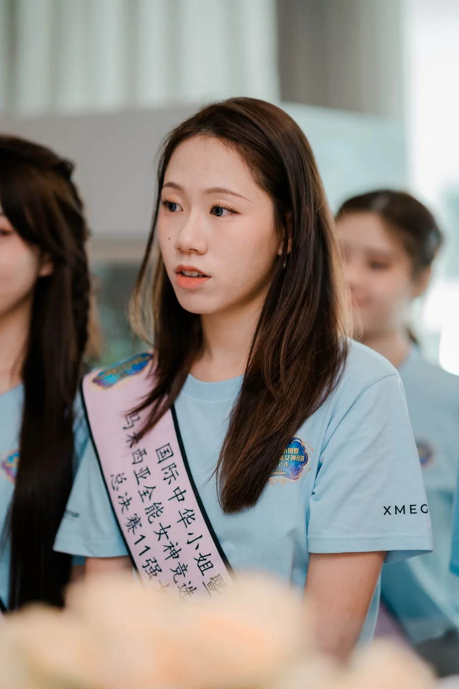 A contestant in a sash and T-shirt listens, seated among other contestants